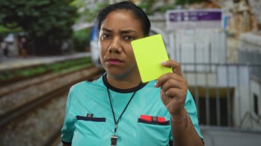 Hakem, kaş çatarken ve turkuaz gömlek giyerken tren istasyonunda sarı kart tutuyor; disiplin.