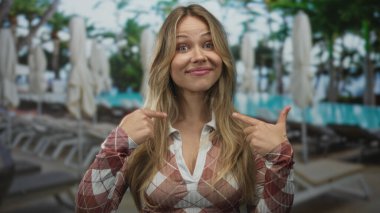 Ekose gömlek giyen gülümseyen sarışın kadın güneşli havuzda şezlongda otururken işaret parmaklarıyla yüzünü gösteriyor..