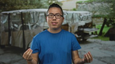 Gözlüklü ve elleri birbirine kenetlenmiş bir adam, arkasında park etmiş arabalarla bir sokakta el kol hareketi yapıyor. İletişim sıkıntısı..