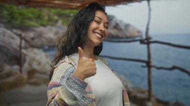 Kadın parmaklarını kaldırır ve kayalık sahilde tahta korkulukların yanında kameraya işaret eder; iyimserlik neşe teşviki.