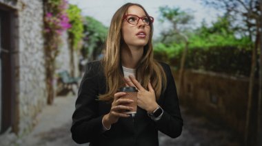 Taş duvar ve yeşilliğin yanındaki bir sokakta elinde kahve fincanı tutan ve kontrol eden kadın; sabırsız işe gidip gelme.