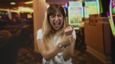 Bir kumarhane binasının içinde bileğini kavrayan ve kumar makinesine bağıran bir kadın, gözleri kapalı ve gergin; hayal kırıklığı.