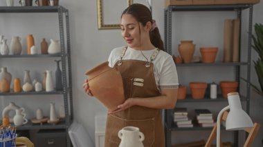 Kadın, çömlek raflarının arasındaki stüdyoda gülümserken iki eliyle kilden bir vazo tutuyor; yaratıcı gurur..