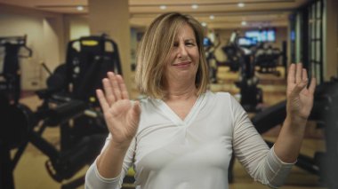 Orta yaşlı beyaz kadın spor salonundaki jestleri durdurmak için egzersiz makineleri arasında el kaldırıyor..