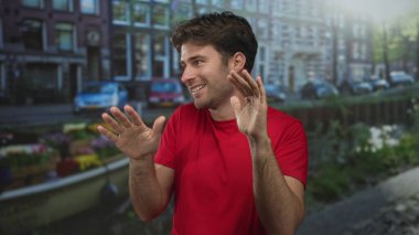 Kırmızı tişörtlü adam, Amsterdam 'da bir kanal caddesinde elleri açık el kol hareketi, önünde avuçlarını hareket ederken gülümseyerek; şakacı rahat.