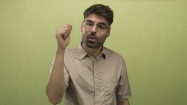 Gözlüklü ve bej renkli kısa kollu genç İspanyol adam yeşil stüdyoda avuç içini gösteriyor..