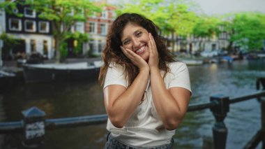 Elleri yanaklarında gülümseyen ve Amsterdam 'da kanal köprüsü üzerinden kollarını kavuşturan bir kadın..