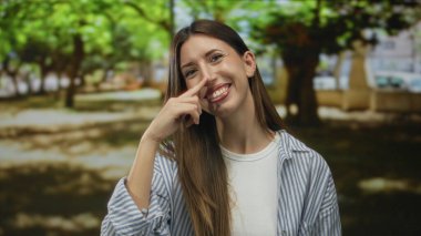 Woman pinches nose under leafy canopy in a sunlit forest filled with bright lush green trees; playfulness.
