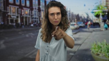 Mavi elbiseli kadın, hareketli caddede sert bir ifadeyle parmağını ileri uzatıyor; meydan okuma güçlendiriyor..