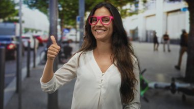 Pembe gözlüklü kadın, arka planda park etmiş scooter 'ı olan işlek bir şehir caddesinde parmağını yukarı kaldırıyor ve baş parmağını kaldırıyor..