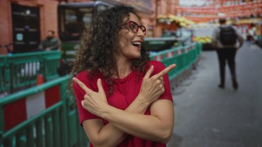 Kıvırcık saçlı, gülümseyen ve işaret eden kırmızı gözlüklü ve gömlekli bir kadın..