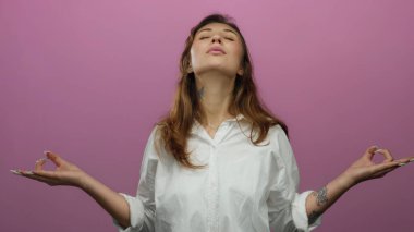 İzole edilmiş pembe arka plana rağmen beyaz gömlekli bir kadın meditasyon yapıyor. Sakince dövmeli sükuneti ve yogayı vurgulayan canlı saçlarıyla sükuneti ve huzuru yayıyor..