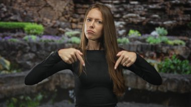 İki işaret parmağı ile kendini işaret eden kadın sokak bahçesinde taş bir duvarın yanında, kaşlarını büzmüş ve kaşlarını kaldırmış;.