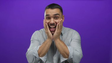 Çizgili gömlekli genç İspanyol adam elleriyle yanaklarına bastırıyor ve mor stüdyoda geniş bir sırıtışla gülümsüyor. Sürpriz..