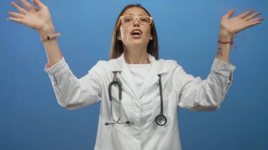 Mavi stüdyoda ellerinde steteskop bardakları olan genç İspanyol kadın doktor eller ağız çevresine; acil halk sağlığı mesajı.