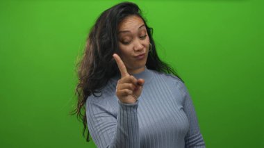 Stüdyodaki kadın yeşil ekranda parmaklarını birbirine gösteriyor, kaşlarını kaldırmış sırıtıyor ve gündelik süveter giyiyor; neşeli bir merak..
