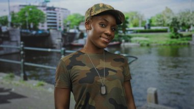 Kamuflaj üniformalı genç bir kadın işlek şehir caddesinde köpek tasması takıyor ve nehir kenarında gülümsüyor..