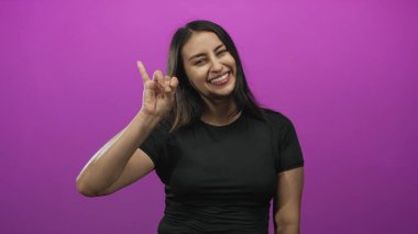 Genç bir kadın gülümsüyor ve mor stüdyoda el hareketi yapıyor. Siyah bir tişört ve görünür bir el ve kol giyiyor..
