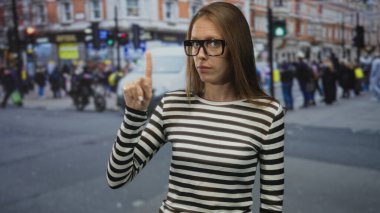 Kadın şehir caddesinde gözlük ve çizgili bluz giyerken sola parmağıyla işaret ediyor; onaylanmıyor.