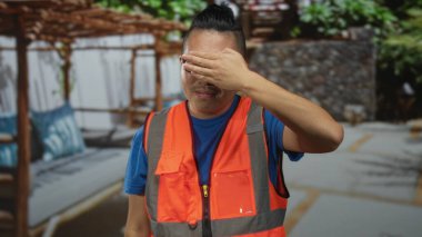 Turuncu yansıtıcı yelek giyen adam, ahşap pergola ve taş duvarın yakınındaki verandada elle gözlerini kapatıyor; çekingenlik belirsizliği.