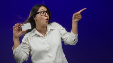 Latin kökenli genç bir kadın beyaz kredi kartı tutuyor ve stüdyoda parmak işareti yapıyor..