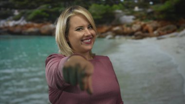 Sarışın kadın, turkuaz su ve kayalık sahil şeridiyle kumlu sahilde kamerayı işaret ediyor..