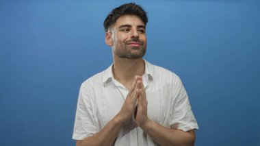 Beyaz keten gömlek giyen İspanyol genç adam mavi stüdyoda el ele tutuşarak dua ediyor..