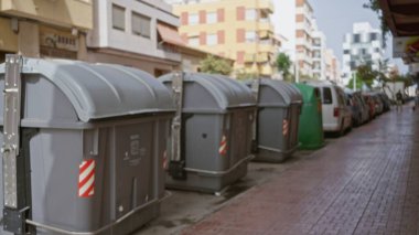 Islak fayanslı bir kaldırım boyunca gri geri dönüşüm kutuları ve park edilmiş kamyonetler, odak binalarından ve ağaçlardan sığ, bulanık sokak arka planına; arka plan arka plan telif alanı sakinliği.