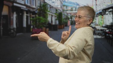 Kadın, diğer el, kısa saç ve gözlük ile gülümseyerek ve göğsüne dokunarak yaya caddesinde ileri doğru işaret eder; neşeli neşe.