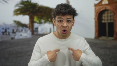 Beyaz kazaklı genç İspanyol adam kilise cephesinin yakınındaki kaldırım taşı sokakta parmak göğse işaret ediyor..