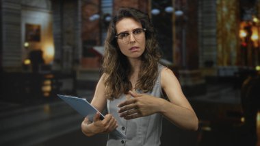 Gözlüklü İspanyol kadın bina içinde kaşlarını çatarken pano ve el hareketleriyle hareket ediyor; belirsizlik kaygısı.