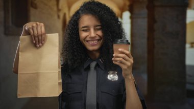 Elinde kahve fincanı ve kağıt torba tutan kadın, eski kasaba sokaklarında gülümsüyor, üniforma giyiyor..