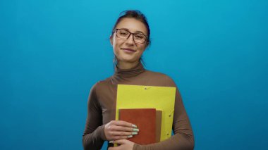 Gülümseyen ve klasörleri tutan bir kadın, izole edilmiş mavi bir arkaplanda, gündelik bir ortamda, kendine güvenini ve tarzını gösteriyor..