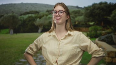 Blonde woman wearing glasses and beige shirt pointing finger to object in forest clearing; discovery curiosity.