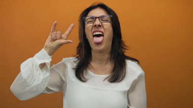 Gözlüklü ve beyaz bluzlu bir kadın stüdyoda parmaklarıyla kaya boynuzları hareketi yapıyor..