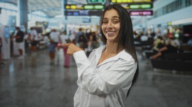 Beyaz tişörtlü genç İspanyol kadın kalabalık havaalanı terminalinde hareket panosunu işaret ederken yandan gülümsüyor ve el kol hareketi yapıyor..