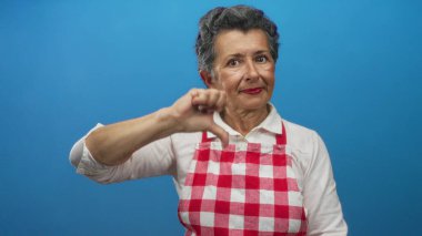 Önlük takan yaşlı bir kadın mavi arka plana karşı olumlu bir jest yapıyor. Ciddi bir ifadeyle memnuniyetsizliğini veya hoşnutsuzluğunu dile getiriyor..