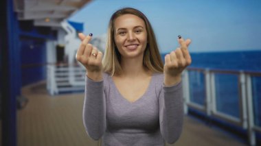 Okyanus manzaralı bir yolcu gemisinde gülümseyen, parmaklarıyla kalp sembolü yapan, uzun kollu bir bluz giyen, barışçıl ve neşeli bir açık hava anı sergileyen bir kadın..