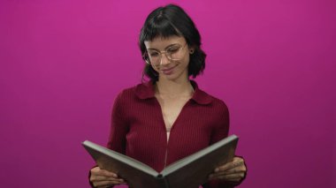Gözlüklü genç İspanyol kadın pembe stüdyo arka planına karşı elleri ile kitaba bakıyor; merak.