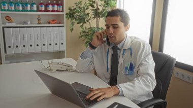 Klinikte steteskobu olan genç İspanyol doktor hastane ortamında dizüstü bilgisayar kullanırken telefonla konuşuyor.