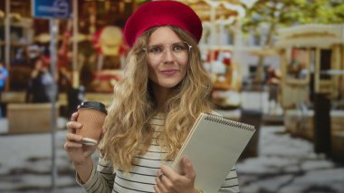 Sokakta elinde kahve tutan, çerçevelenmiş sarı saçlarıyla eskiz defterini gösteren ve şehirde gözlüklü bir berenin altında gülümseyen bir kadın..