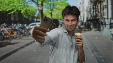 Adam şehrin caddesinde akıllı telefon tutarken dondurma külahıyla selfie çekiyor; neşeli yaz anı.