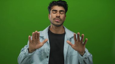 Ciddi ifadeli kot gömlek giyen İspanyol genç adam yeşil stüdyoda avuçlarını ileri doğru tutar; sakin ol..