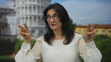 Orta yaşlı, gözlüklü İspanyol kadın, dışarıdaki işeme kulesinin yanında parmaklarıyla havadan tırnak işareti yapıyor..