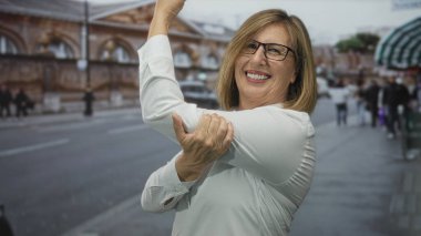 Beyaz gömlekli orta yaşlı beyaz kadın şehir caddesinde bulanık binaları ve insanları geçerek kolunu esnetiyor; güven güçlendiriyor.