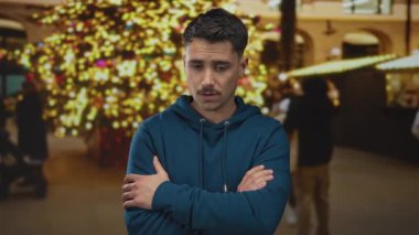 Çapraz kollu genç İspanyol bir adam, canlı bir akşamda düşünceli bir ruh hali somutlaştıran, parlak tatil ışıklarının şenlikli bir arka planına karşı bir şehir caddesinde duruyor..