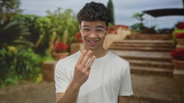 Gözlüklü ve beyaz gömlekli adam, parkta taş basamaklar ve yemyeşil alanda yumruklarını sıkıyor; heyecan.