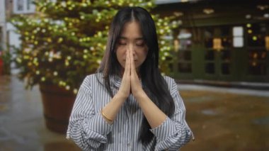 Dışarıda dua ederken ellerini sımsıkı bağlayan genç bir kadın hayat dolu bir caddede süslü bir Noel ağacının önünde duruyor huzur ve sükuneti ifade ediyor..