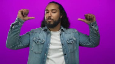 Mor stüdyoda göğsünü gösteren başparmaklı bir adam kot ceket ve beyaz tişört giyiyor..