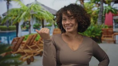 Woman smiles with hand on hip under palm trees by sunlit poolside at lush tropical resort; vacation escape serenity.